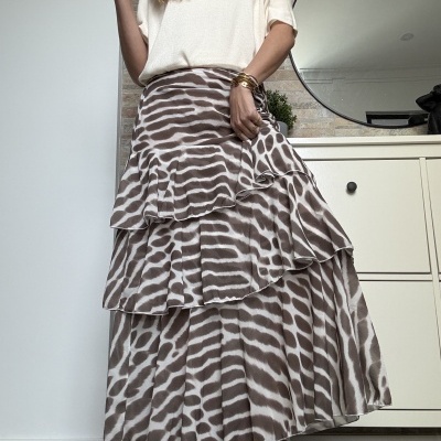 Saia comprida com padrão castanho e branco estilo zebra com folhos, t-shirt branca, pulseiras douradas e sapatilhas castanhas e brancas numa divisão com móvel branco e espelho