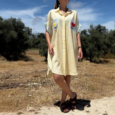 Vestido curto amarelo com riscas brancas e aplicações de sol e coração em bolso, mulher de sapatos pretos, ao ar livre com árvores e céu azul