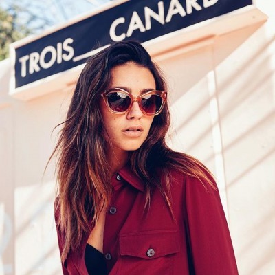 Mulher com óculos de sol castanhos e camisa vermelha junto a placa 'TROIS CANARDS'