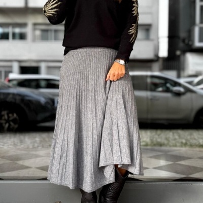Mulher com saia plissada cinzenta, camisola preta com bordados dourados e botas pretas em ambiente urbano