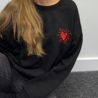 Pessoa a usar sweatshirt preta com coração vermelho bordado no peito.