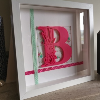 Quadro com letra decorativa B rosa com fitas coloridas em moldura branca