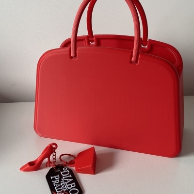 Mala vermelha em silicone com acessórios vermelhos e etiqueta preta com texto O DIABO VESTE PRADA.