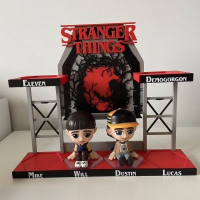 Display acrílico com figuras Stranger Things Mike, Will, Dustin e Lucas, com texto vermelho e fundo preto e vermelho.