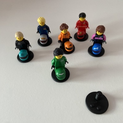 Seis figuras LEGO coloridas em bases pretas sobre superfície branca com peça preta solta ao lado