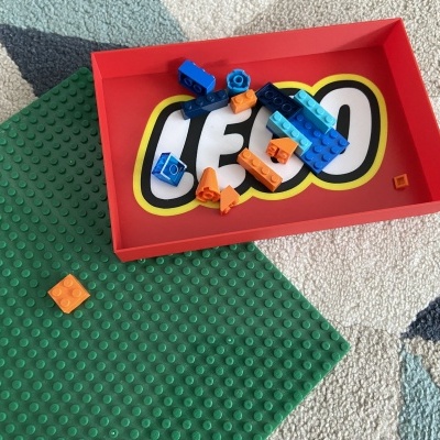 Peças LEGO coloridas numa caixa vermelha com logo LEGO, sobre tapete e base de construção verde.