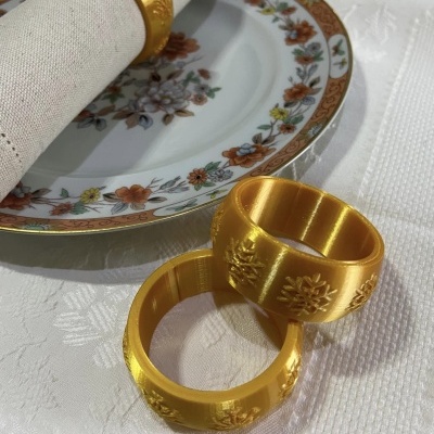 Anéis porta-guardanapos dourados com padrão floral ao lado de prato decorado