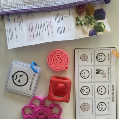 Objetos coloridos de plástico, cartões com expressões faciais e texto em português, folheto e bolsa de malha roxa sobre mesa clara.