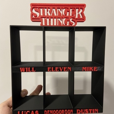 Estante preta com nomes em relevo vermelhos e texto STRANGER THINGS no topo