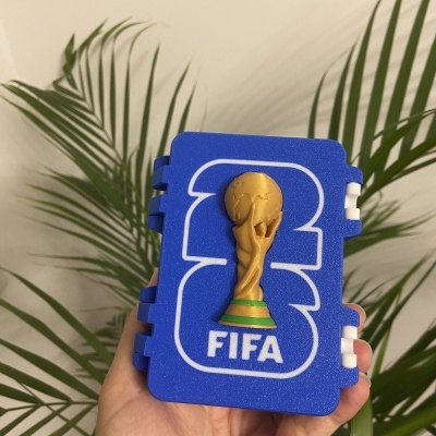Caixa azul com logotipo da FIFA e taça do mundo em relevo dourado segura por mão
