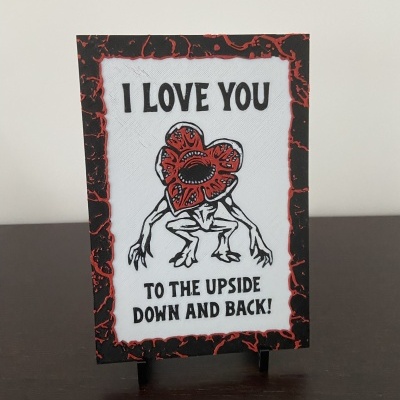 Placa decorativa com texto I LOVE YOU TO THE UPSIDE DOWN AND BACK e figura de criatura com cabeça em forma de flor vermelha.