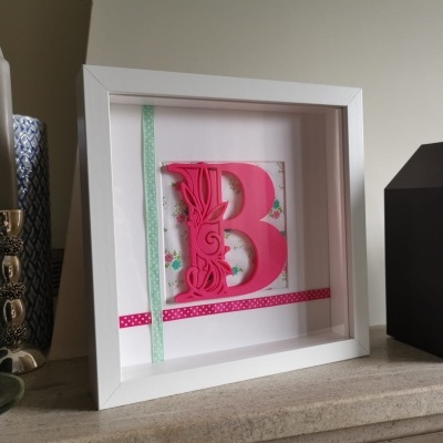 Quadro decorativo com letra B rosa e moldura branca junto a castiçal, vaso azul e relógio preto