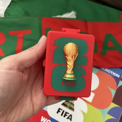 Caixa vermelha com logotipo FIFA e troféu dourado, mão segurando, pano verde e álbum FIFA 2022 ao fundo