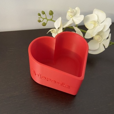 Recipiente vermelho em forma de coração com palavra 'Adorarte' e flor branca ao fundo