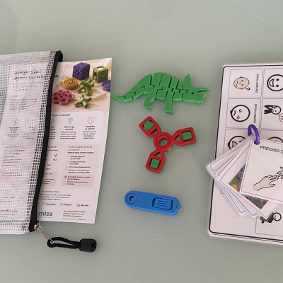 Kit de brinquedos antistress com bolsa, folhetos explicativos e cartões comunicativos em superfície cinza.