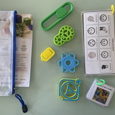 Kit de brinquedos fidget coloridos com bolsa transparente e folhas informativas em português sobre superfície cinza