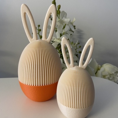 Dois coelhos decorativos creme com base laranja e branca em superfície branca e flores ao fundo