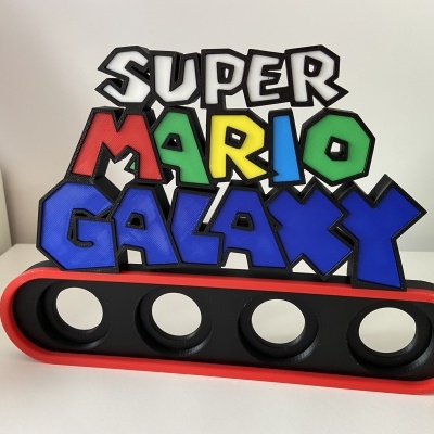 Objeto decorativo com as palavras SUPER MARIO GALAXY em várias cores sobre base preta e vermelha