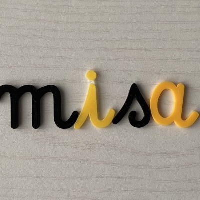 Palavra decorativa 'misa' em plástico preto e amarelo sobre superfície clara de madeira