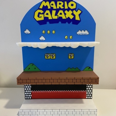 Expositor Super Mario Galaxy com fundo azul, letras amarelas, nuvens, moedas, tijolos castanhos e base tijolada