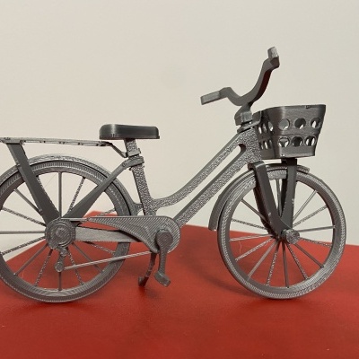 Miniatura de bicicleta cinza metálica com cesta e suporte, sobre superfície vermelha.