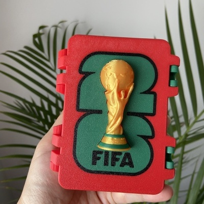 Caixa vermelha com relevo dourado da Taça do Mundo FIFA e palavra FIFA, mão segurando, planta ao fundo.