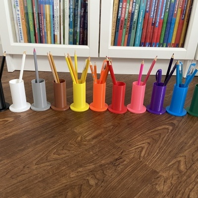 Onze suportes coloridos para lápis com lápis dentro, em chão castanho, móveis e livros ao fundo.