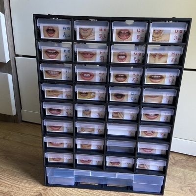 Organizador preto com gavetas transparentes contendo imagens de bocas e letras, sobre chão de madeira.