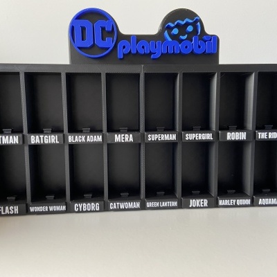Expositor preto vazio com múltiplos compartimentos etiquetados com nomes de personagens DC e logótipo DC Playmobil azul no topo.