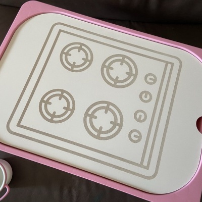 Tábua de cortar com desenho de fogão e suporte rosa sobre sofá castanho com chávena branca