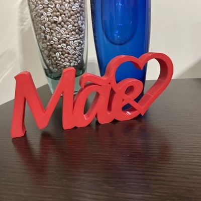 Figura decorativa vermelha da palavra 'Mãe' com coração numa superfície de madeira escura ao lado de vasos