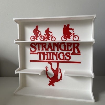 Estante branca de parede com decoração vermelha Stranger Things e silhuetas de crianças e figura pendurada