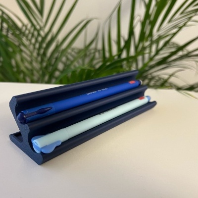 Suporte dobrável azul com duas canetas, uma azul e outra branca, em fundo branco com planta verde