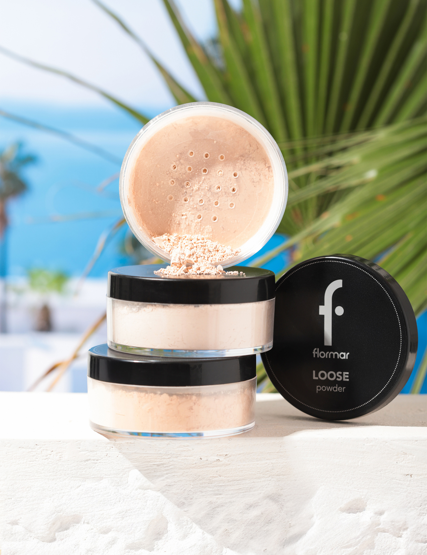 Frascos de pó solto da marca flormar com tampa preta e pó bege, com palmeira e mar ao fundo.
