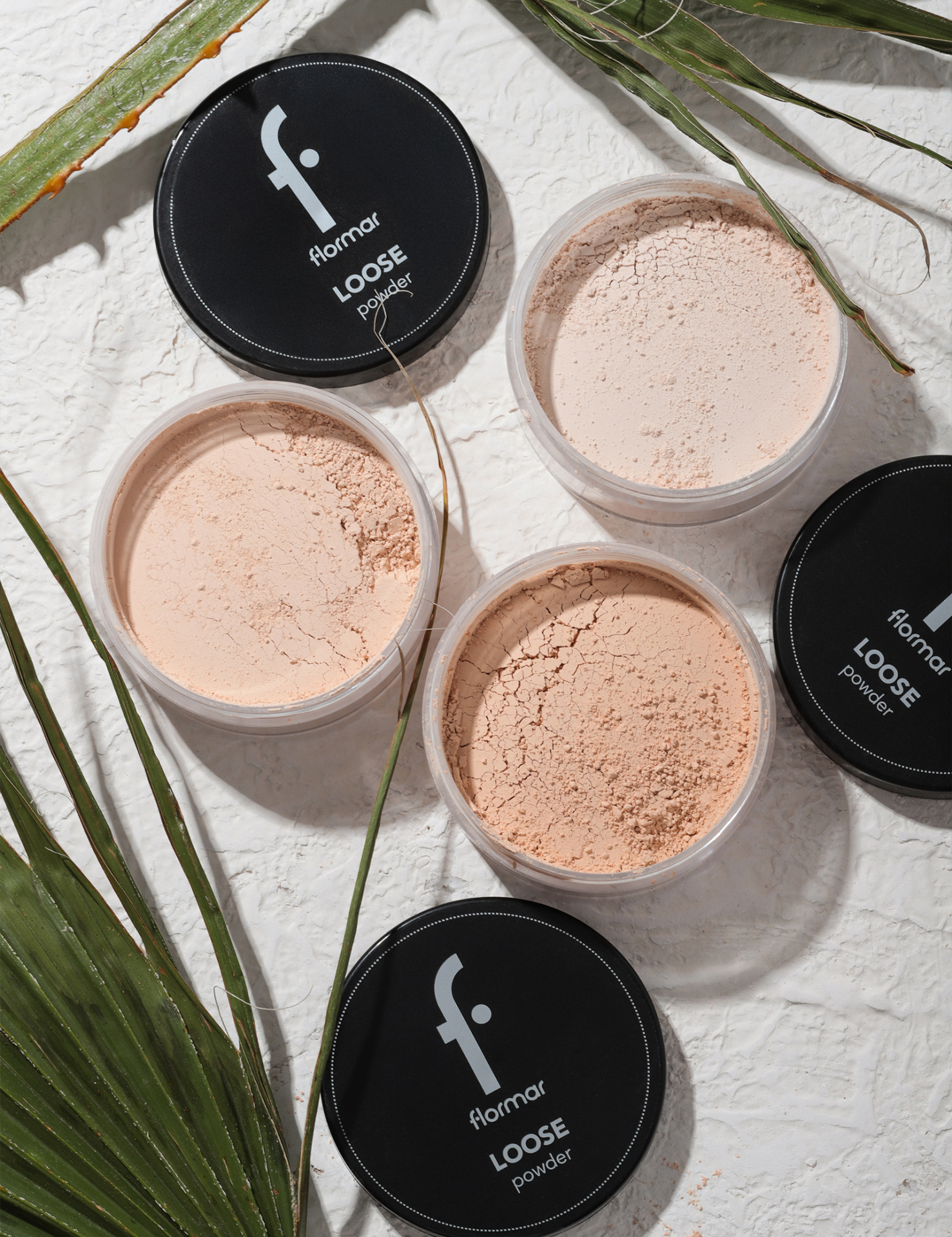Pós solto Flormar em recipientes transparentes com tampa preta e rótulo visível, sobre superfície branca com folhas verdes.