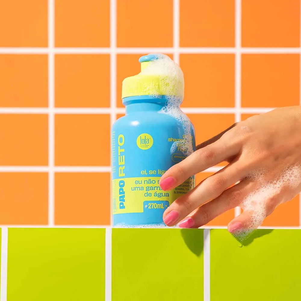 Garrafa azul com tampa amarela e espuma branca, texto em amarelo e azul, mão segurando, fundo de azulejos laranja e verde