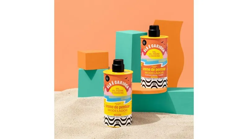 Dois frascos amarelos de creme de pentear com rótulos coloridos sobre areia e blocos coloridos ao fundo