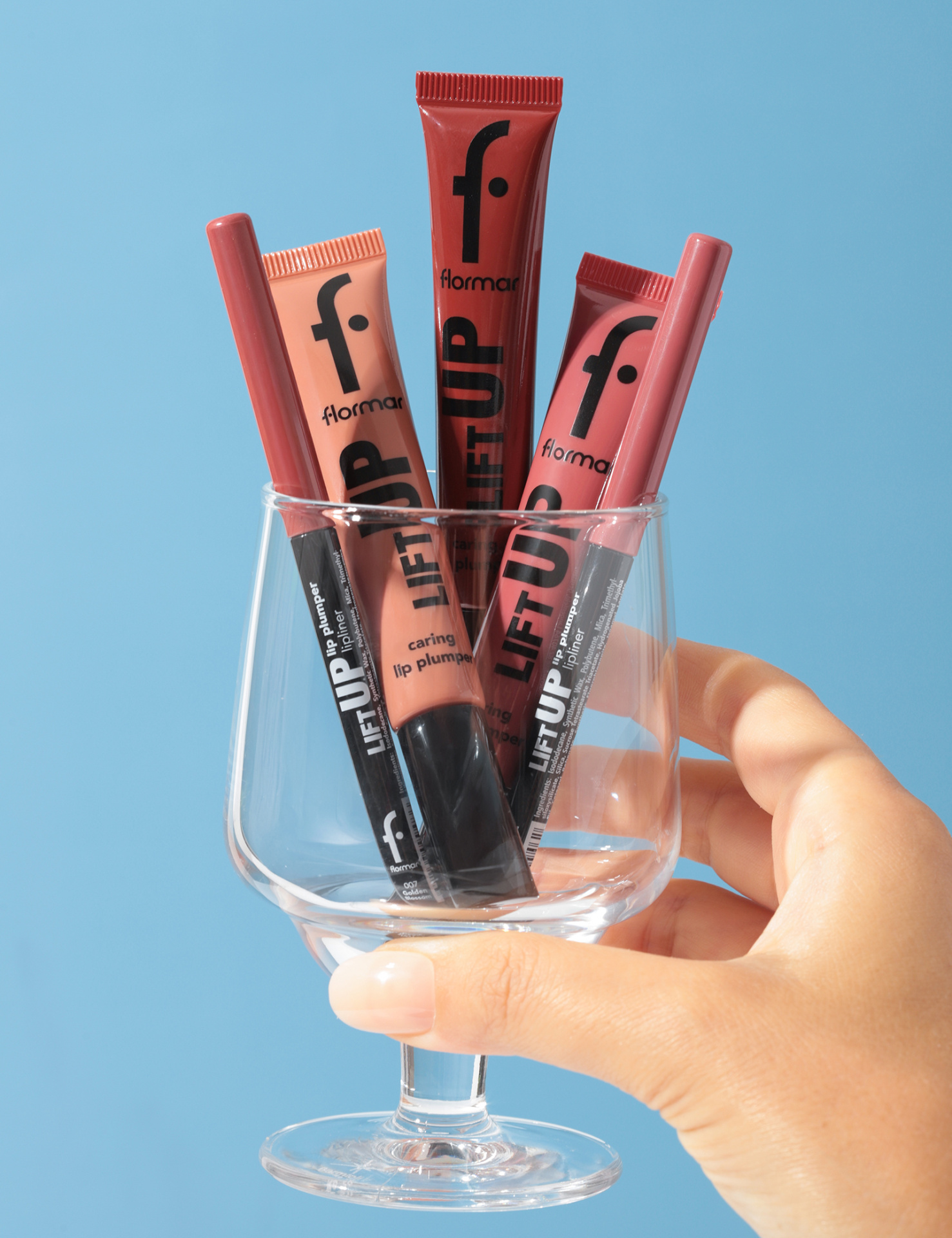 Tubos de batom Flormar em copo transparente segurado por mão feminina, fundo azul claro.