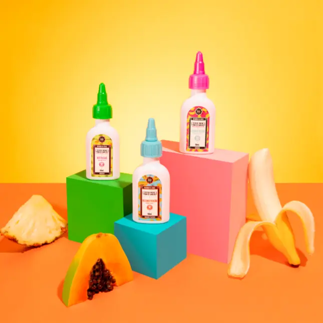 Três frascos de plástico branco com tampas coloridas sobre cubos de madeira e frutas tropicais em fundo colorido