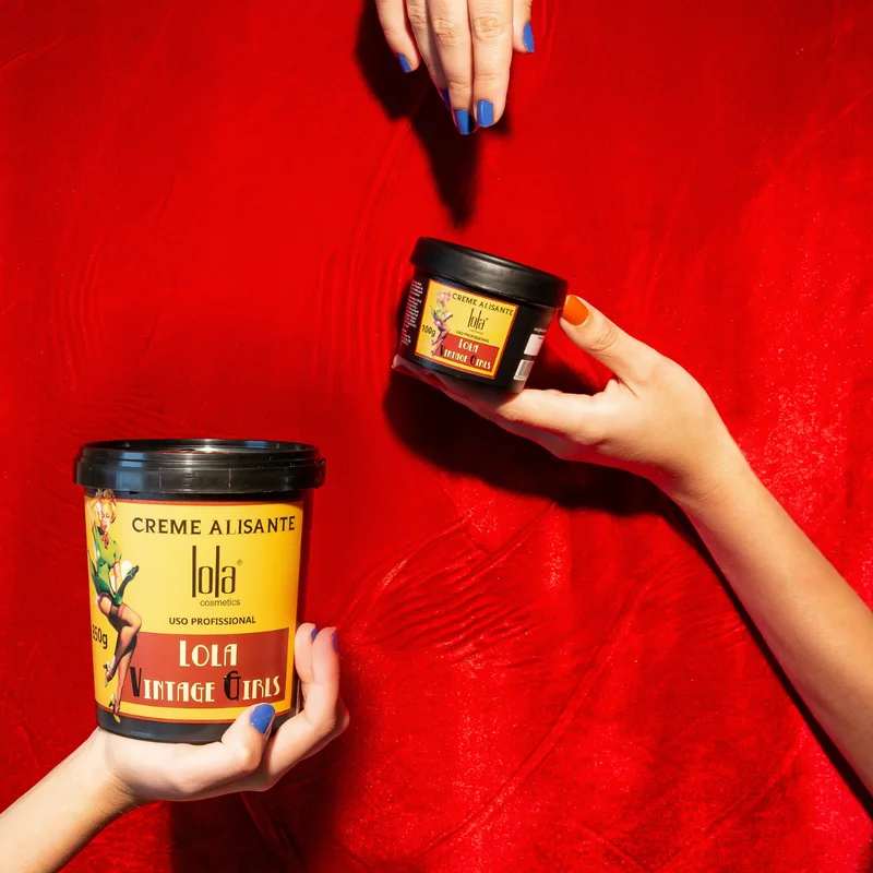Dois frascos de creme alisante Lola Vintage Girls em fundo vermelho, mãos segurando os frascos.