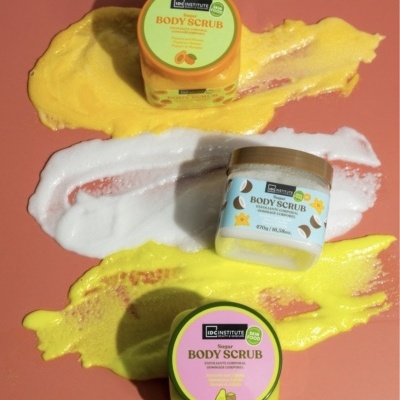 Três frascos coloridos de body scrub sobre fundo rosa com amostras de creme amarelo e branco