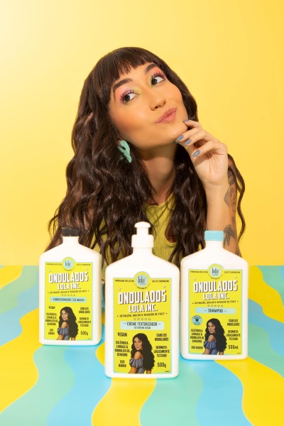 Três produtos de cuidados capilares Ondulados Lola Inc. com rótulos amarelos sobre mesa colorida, mulher jovem ao fundo