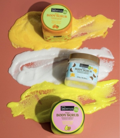 Três frascos coloridos de body scrub sobre fundo rosa com amostras de creme amarelo e branco