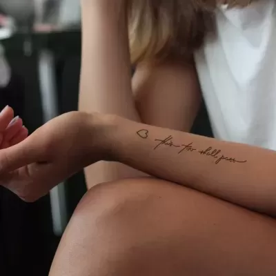 Braço feminino com tatuagem de coração e texto 'faith-hope-dream' em caligrafia preta