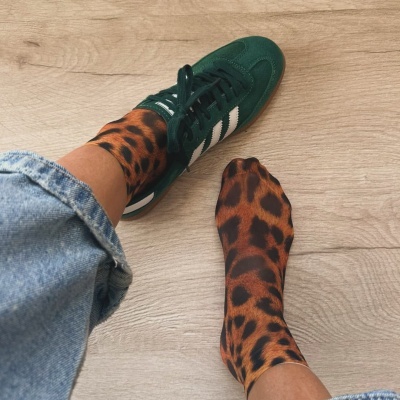 Meias com estampa animal castanha e preta, calças de ganga azuis e sapato desportivo verde sobre chão de madeira.