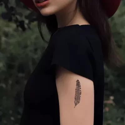 Braço com tatuagem de pena preta em pele clara, camiseta preta de manga curta, fundo de vegetação desfocada