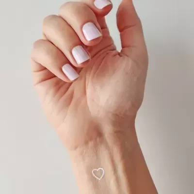 Mão com unhas pintadas de rosa claro e tatuagem de coração no pulso