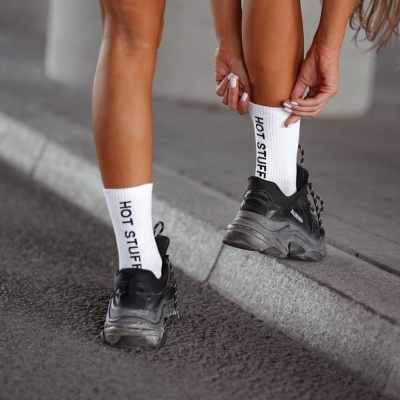 Meias brancas com texto 'HOT STUFF' usadas com sapatilhas pretas numa rua asfaltada
