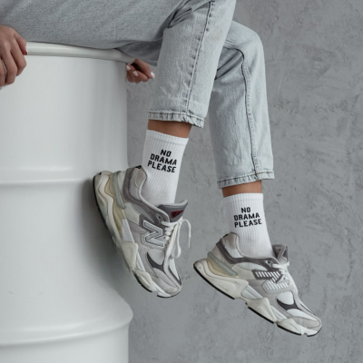 Meias brancas com texto e sapatilhas cinza com detalhes, calças de ganga claras, sentada num barril branco