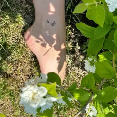 Pé com tatuagens pequenas perto de flores brancas e folhas verdes